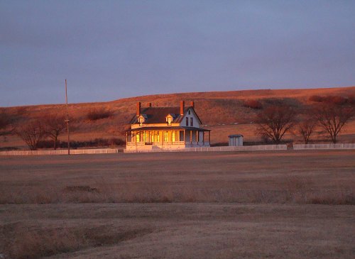 A historic house bathed in golden sunset light, surrounded by open fields and gentle hills.