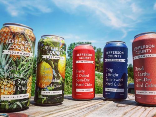 Five colorful cans of Jefferson County Ciderworks hard cider on a wooden table, set against a blue sky.