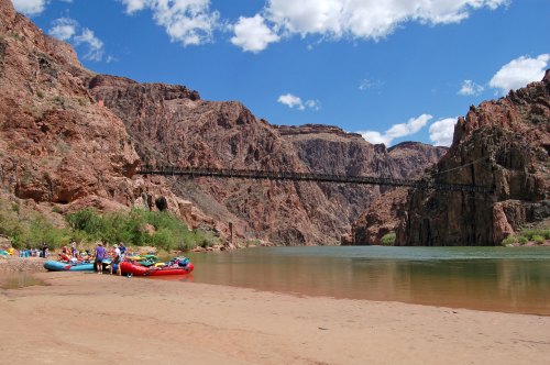 A scenic view of a river with rafts on the shore, surrounded by rocky cliffs and a bridge in the background.