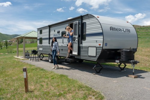 Two women stand outside a travel trailer in a grassy area, with a blue sky and hills in the background.