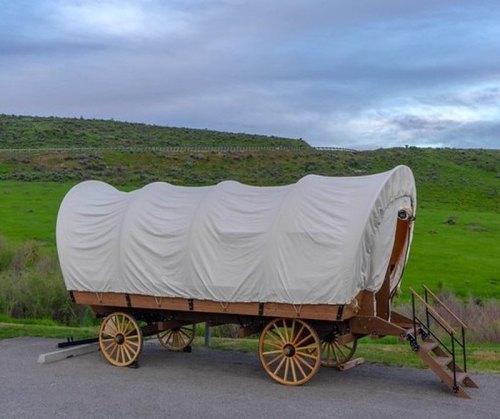 A covered wagon with a white canvas top, parked on a road with green hills in the background under a cloudy sky.