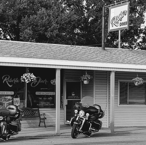 Black and white image of Roberta's Diner with two motorcycles parked outside. Trees in the background.
