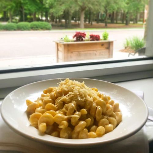 A bowl of creamy pasta topped with grated cheese, set against a window view of greenery and a wooden planter.