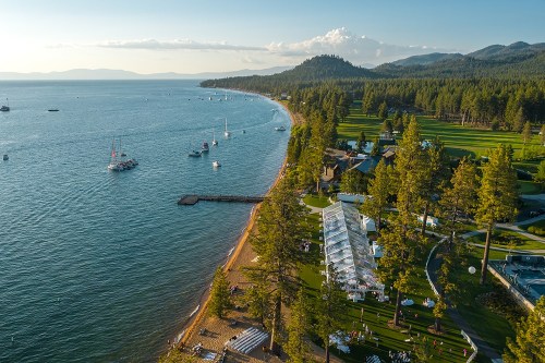 Aerial view of a lakeside venue with boats, sandy beach, and lush green trees under a clear sky.