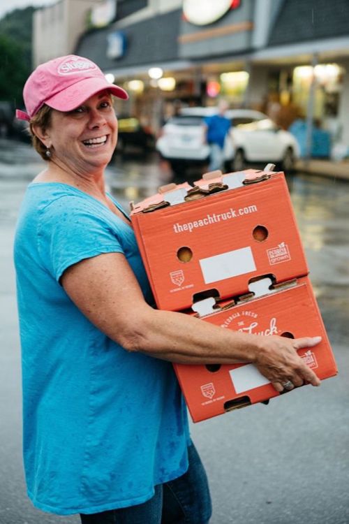 A smiling woman in a pink cap carries two orange boxes labeled "thepeachtruck" on a rainy street.