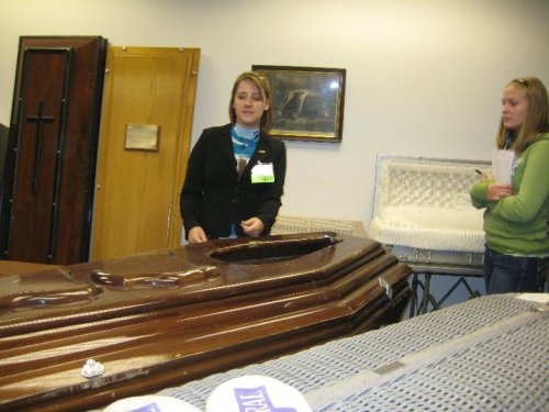 Two women in a room with various coffins, one speaking while the other listens, with a serious atmosphere.