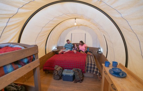 A family sits on a bed in a cozy, tent-like room with wooden furniture and camping gear.