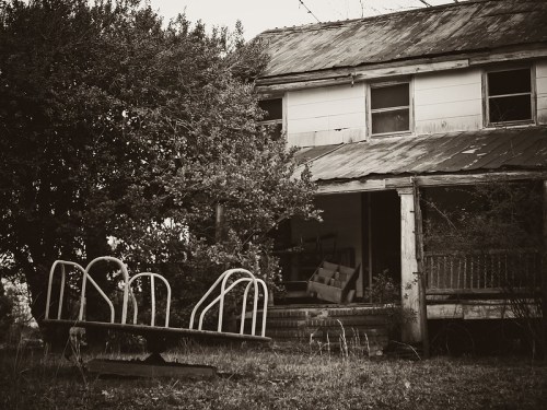An abandoned house with a rusty playground structure in the foreground, surrounded by overgrown vegetation.
