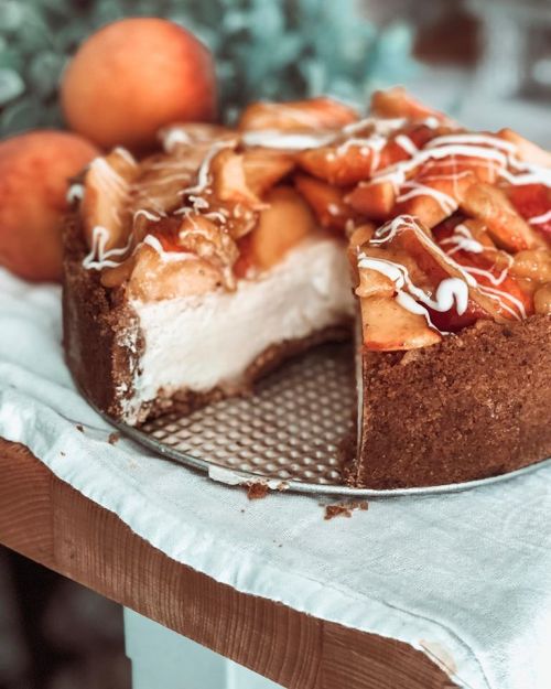 A delicious cheesecake topped with caramelized apples, drizzled with icing, on a wooden table with fresh peaches nearby.