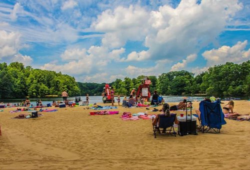 A sandy beach scene with people sunbathing, trees in the background, and water slides nearby under a blue sky with clouds.