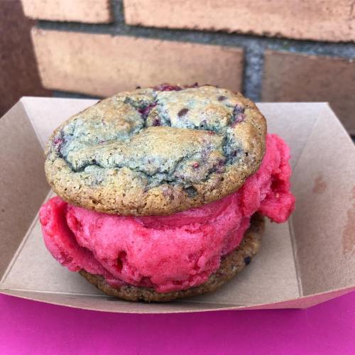 A colorful ice cream sandwich with pink sorbet between two chocolate chip cookies, served in a paper tray.