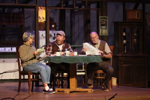Three actors sit around a table in a cozy set, reading scripts and engaging in conversation.
