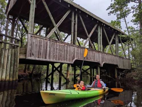 A child and adult kayak under a wooden bridge in a serene, wooded area.