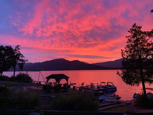 A vibrant sunset paints the sky in pink and purple over a calm lake, with silhouettes of trees and boats in the foreground.