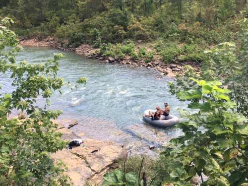 Two people relax on an inner tube in a river, surrounded by lush greenery and rocky banks.