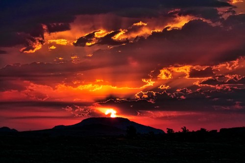 A vibrant sunset casts orange and red hues over a silhouetted mountain, with dramatic clouds in the sky.
