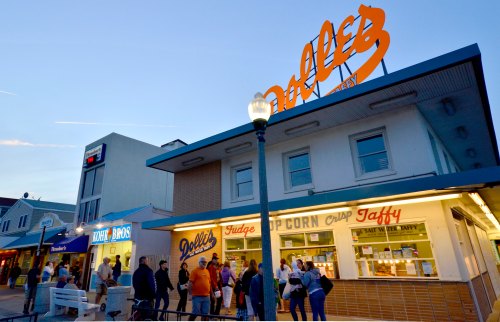 A bustling boardwalk scene at dusk, featuring a candy shop with a bright sign and a crowd of people outside.