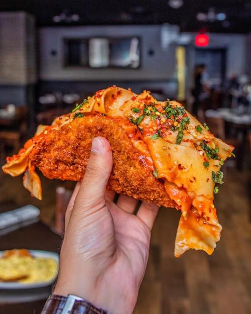 A hand holding a crispy taco filled with pasta and sauce, garnished with herbs, in a restaurant setting.