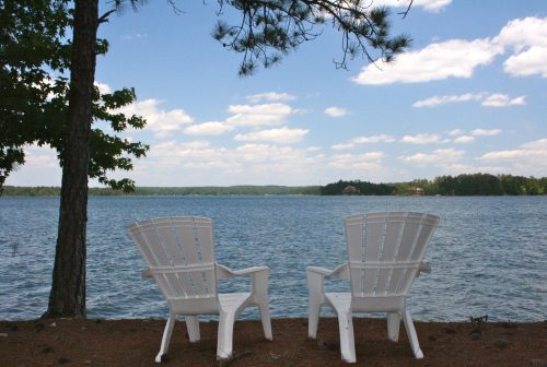 Two white chairs facing a serene lake under a blue sky with fluffy clouds, surrounded by trees.