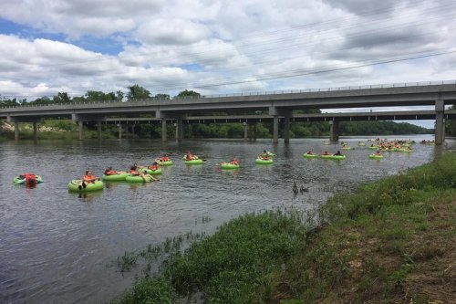 A group of people in green inflatable rafts float down a river under a bridge, with cloudy skies above.