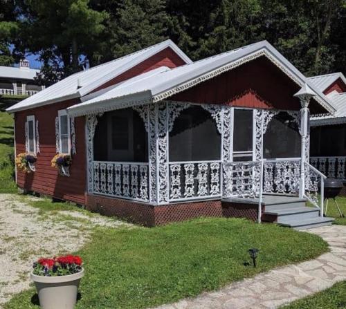 A charming red cottage with white decorative trim, surrounded by greenery and flowers, featuring a porch and a grill.