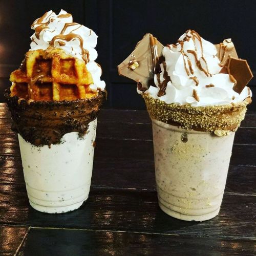 Two decadent milkshakes topped with whipped cream, chocolate drizzle, and waffle pieces, served in unique cone cups.