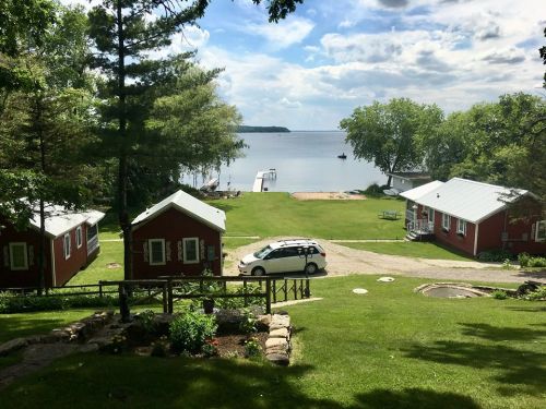A scenic view of a lake with red cabins, green grass, and a car parked on a gravel path under a blue sky.
