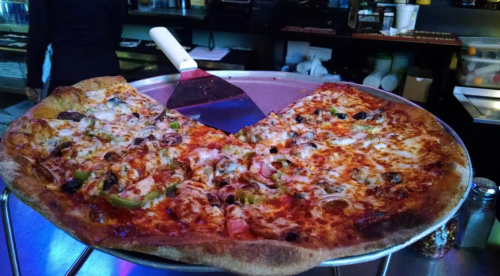 A large pizza on a serving tray, topped with cheese, vegetables, and meats, with a pizza cutter resting on top.