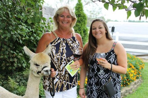 Two women smile while holding drinks, standing next to an alpaca in a garden with flowers and greenery.