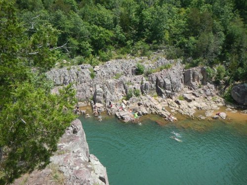 A scenic view of a rocky shoreline with people swimming and relaxing in a clear green lake surrounded by trees.