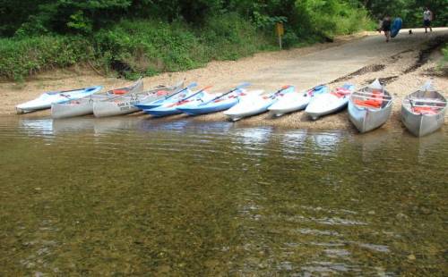 A row of canoes and kayaks lined up on a pebbly shore beside a calm river, with people walking in the background.