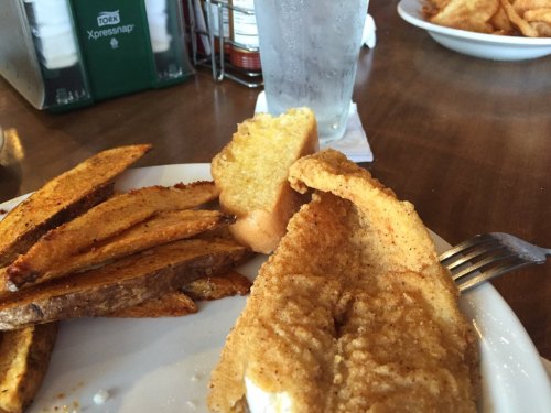A plate with fried fish, golden-brown potato wedges, and a slice of bread, alongside a glass of water.
