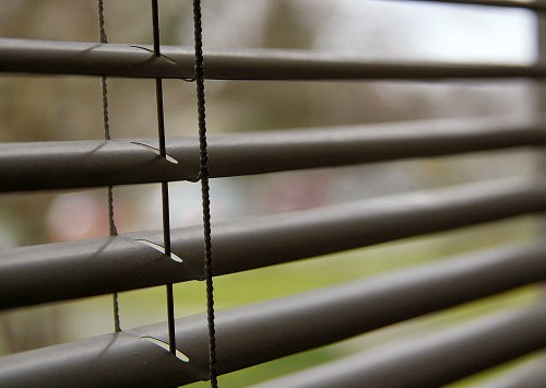 Close-up of horizontal blinds with soft light filtering through, casting shadows on a blurred outdoor background.