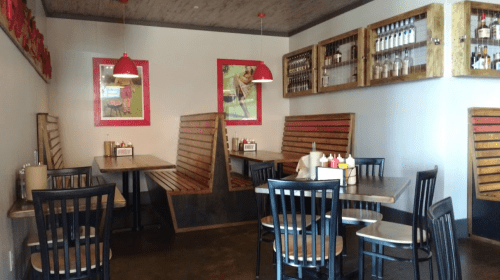 Cozy restaurant interior with wooden booths, tables, and red pendant lights, featuring colorful wall art and a drink display.