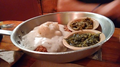 A metal dish with fried chicken smothered in gravy, topped with an egg, and two sides of greens.