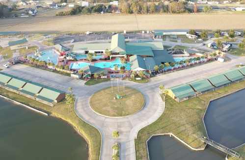 Aerial view of a resort with pools, slides, and surrounding greenery, near a lake and open fields.