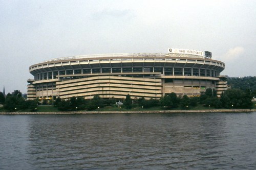 A large stadium viewed from across a river, surrounded by trees and a cloudy sky.