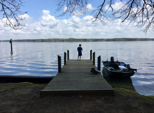 A person stands on a wooden dock by a calm lake, with a small boat nearby and trees in the background.