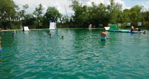 A lively swimming area with people enjoying the water, inflatable slides, and trees in the background under a blue sky.