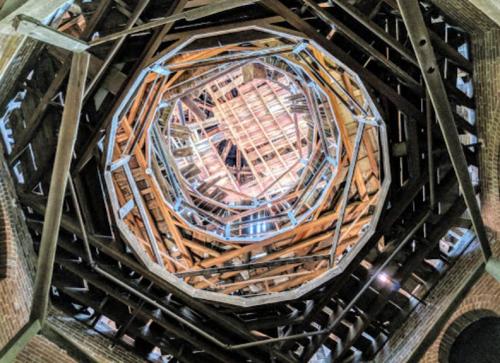 View looking up at a circular wooden structure with intricate beams and a bright light at the center.
