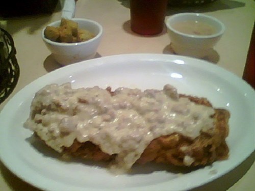 A plate of fried chicken smothered in creamy gravy, with a side of okra and a small bowl of soup.