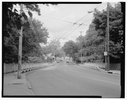 A quiet street scene with trees lining the road, power lines overhead, and a bridge in the distance under a cloudy sky.
