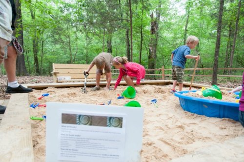 Children playing in a sandy area with toys, surrounded by trees and a sign about the activity.