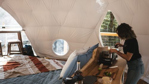 A cozy geodesic dome interior with a woman cooking at a wooden table, featuring a bed and large circular window.