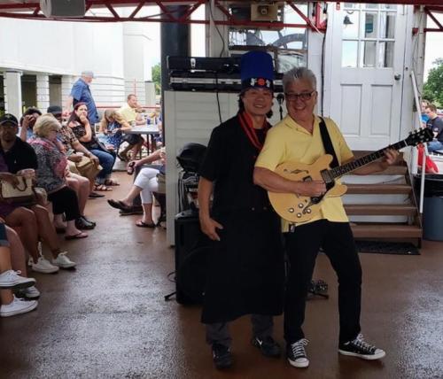 Two musicians pose together on stage, one in a chef's hat and apron, the other playing guitar, with an audience in the background.