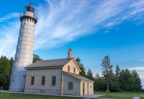 A tall, silver lighthouse beside a stone house, set against a clear blue sky and green trees.