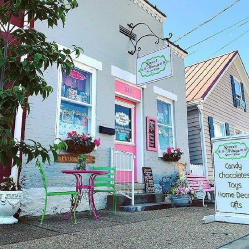 Charming storefront of "Sweet Things" with colorful decor, flowers, and a sign for candy, toys, and gifts.