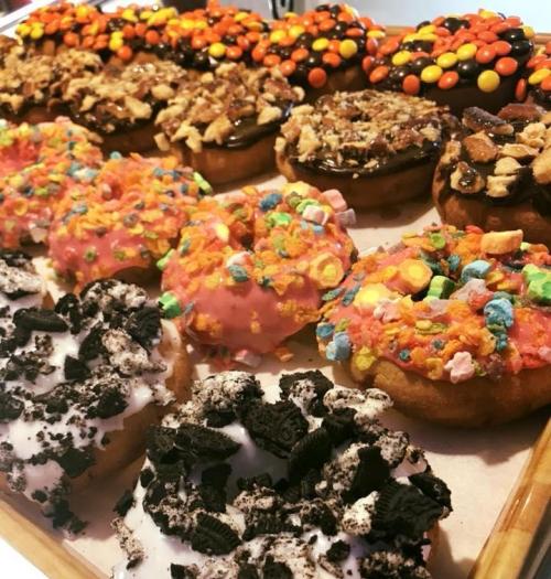 A variety of colorful donuts topped with candy, chocolate, and crushed cookies, arranged on a wooden tray.