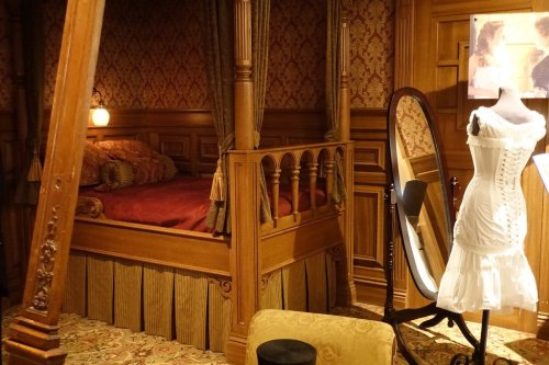 A vintage bedroom featuring a wooden four-poster bed, a mirror, and a white dress on a mannequin.