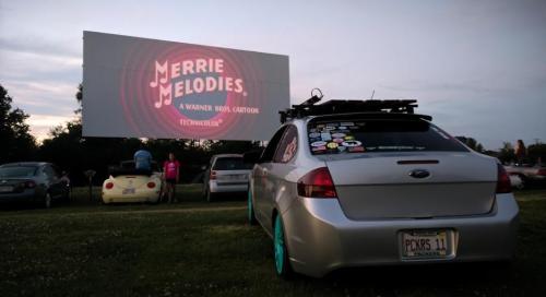A car parked at a drive-in theater with a "Merrie Melodies" screen in the background during twilight.